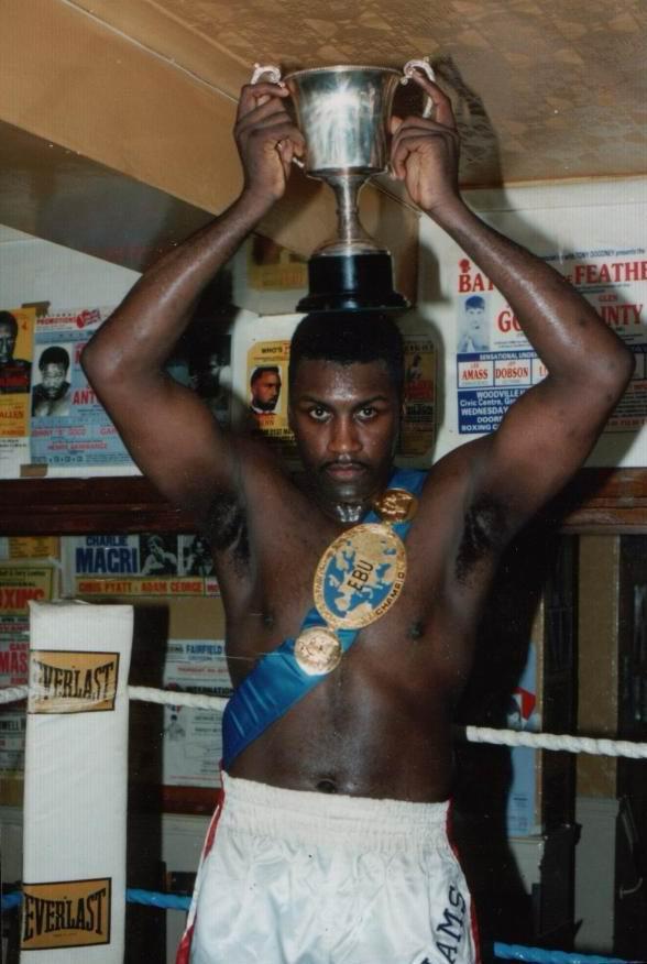 A young Derek holding a trophy aloft in the gym, wearing a championship belt